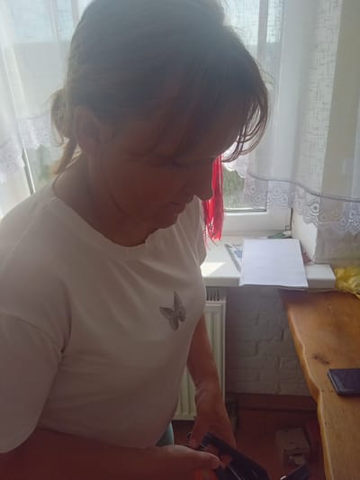 A woman with brown hair tied back is looking down and to her right. She is wearing a white t-shirt with a silver butterfly emblem on the left side of her chest. She is standing in front of a window with white curtains. To her right, on a wooden surface, are a spiral notebook, a yellow plastic bag, and a smartphone. The environment appears to be indoors, possibly a residential setting, given the window, curtains, and radiator visible in the background.