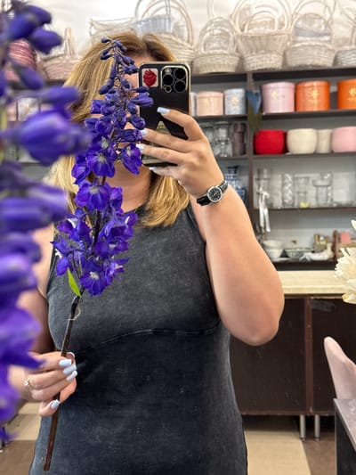 The media file is a selfie taken in a room filled with shelves displaying various items. The primary subject is a person, whose arm and hand are visible, holding a smartphone. The person is wearing a dark-colored, ribbed top and a watch with a black band and a silver face on their left wrist. Their nails are painted a light blue color. The person is holding a stem with several vibrant purple delphinium flowers. The background shows shelves stocked with items like woven baskets, cylindrical containers in colors such as orange, pink, and red, and other unidentifiable objects. The lighting suggests it is daytime, and the setting appears to be an indoor retail space or storage area, possibly a flower shop or craft store, given the presence of flowers and baskets. The overall mood is casual and personal, typical of a selfie. There is no visible text in the image.