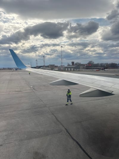 The image depicts an airport scene in Kitsjanovo, Russia, viewed from an aircraft window looking down onto the tarmac. A large white airplane wing, with a distinctive blue wingtip, spans the lower half of the frame. The tarmac is dark asphalt, showing various painted lines in red and white, along with some dark stains or tire marks.

A ground crew member is prominently visible on the tarmac next to the wing, wearing a high-visibility yellow-green jacket with black elements, black trousers, and black shoes with yellow-green accents. This individual appears to be walking or standing as part of the airport's ground operations. Another, less distinct, crew member can be seen further down the wing towards the background.

The background features several buildings, possibly airport infrastructure or hangars, behind which a line of bare trees suggests a colder season. Utility poles or antennae also rise into the sky. Two national flags are visible on poles; one is clearly the red flag with a white cross of Switzerland, and next to it, a red and white horizontally striped flag is partially visible.

The scene is set during the daytime under a heavily overcast sky, characterized by dramatic formations of grey and white clouds. The overall impression is one of typical airport activity, with an aircraft parked on the apron and ground crew present. No discernible text is visible on any objects within the image.
