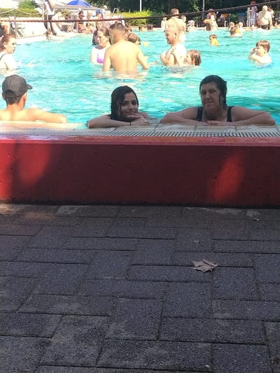 The image shows a group of people in a swimming pool on a sunny day. The subjects include men, women, and children, many of whom are swimming or relaxing in the water. One woman is leaning on the edge of the pool, smiling at the camera. Another person, a child, is partially submerged in the water, with their head and shoulders visible. The pool is surrounded by a tiled area, and in the background, there are trees and other vegetation, suggesting an outdoor location. The overall mood of the image is relaxed and enjoyable, with people taking advantage of the warm weather to swim and socialize. The lighting suggests it is daytime, and the presence of people in swim attire indicates a recreational activity. No visible text is present in the image. The location is Herford, Germany.