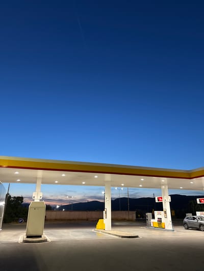 The image depicts a gas station in Kalotina, Bulgaria, identified by the Shell logo and signage. A gray car is parked near pump number 6. The sky is a deep blue. Behind the pumps, there is a fence, trees, mountains, and the setting sun.