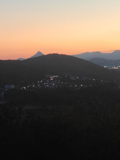 The image shows a twilight view of a hillside settlement in Međugorje, Bosnia and Herzegovina. The sky is a gradient of pale blue to soft orange, indicative of sunset or sunrise. In the foreground, the dark, undulating slopes of hills are covered in vegetation. Scattered across the hillside are lights from buildings, suggesting a small community. One cluster of lights appears to be concentrated in a village. In the distance, a prominent, sharply peaked mountain dominates the skyline, with other fainter mountain ranges visible beyond it. The overall impression is one of a rural landscape illuminated by the warm glow of evening or morning light and the man-made lights of the settlement.