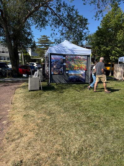 In the Village of Sag Harbor, United States of America, on a sunny day, a white tent displays art for sale. Two people, a man and a woman, walk past the art display. The man is wearing a dark t-shirt and khaki shorts, while the woman is wearing a white shirt and blue jeans. Behind the tent, vehicles are parked, including a red car and a black SUV. The art tent is set up on a grassy area adjacent to a gravel path. Trees with green leaves are visible in the background, and a clear blue sky completes the scene.
