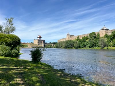 This is a wide-angle, eye-level outdoor shot of the Ivangorod Fortress in Ivangorod, Russia, on a bright, sunny day. The fortress, a prominent stone structure with several towers, stands on the far bank of a wide, calm river. The river occupies the majority of the foreground and middle ground, with ripples on its surface reflecting the blue sky. Lush green trees and bushes line the far bank of the river, creating a dense, natural barrier between the water and the fortress walls.
On the near side of the river, a grassy bank slopes down to the water's edge. The grass is a vibrant green, dappled with sunlight and shadows, suggesting it's late morning or early afternoon. A few small, dark green bushes are scattered on this bank. The sky above is a clear, bright blue, streaked with wispy white clouds that add texture and depth.
The Ivangorod Fortress itself is the main subject, appearing as a formidable and historic landmark. Its stone walls are weathered, and the towers, topped with red-tiled roofs, suggest a long history. In the distance, behind the fortress, other buildings and spires are visible, hinting at a town or city beyond. The overall scene conveys a sense of tranquility and historical significance, with the natural beauty of the river and trees contrasting with the man-made architecture of the fortress. There is no visible text in the image.