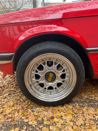 The image shows the front wheel of a red car parked on a surface covered with fallen autumn leaves. The wheel is a silver BBS design with gold-colored bolts. The tire is a Mozzo 4S+. The location is inferred to be somewhere in the city of Florida, Argentina, due to the presence of autumn leaves indicating the presence of deciduous trees and the car being in the region of Argentina. No people are visible.