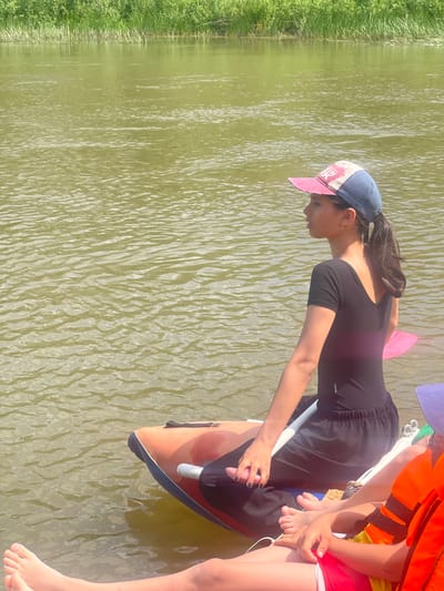 A young person wearing a black one-piece bathing suit and a baseball cap sits on the edge of a colorful inflatable raft in a river. The raft is orange with blue trim. The person is looking to the right, with a paddle resting on their lap. The river is murky green and reflects the bright sky. In the background, a lush green embankment lined with tall grasses borders the river. Another person, wearing an orange life vest and a purple hat, is also visible on the raft. The scene appears to be a warm, sunny day. The city of Nezhinka, Russia is not depicted in this image.