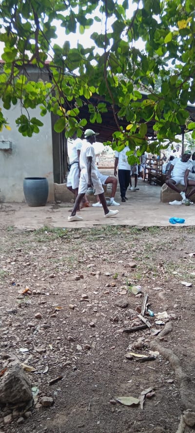 A group of men dressed in white uniforms are gathered in an outdoor setting. Some are walking, while others are seated. One man carries what appears to be a kettlebell. The scene is partially framed by tree branches with green leaves. The ground in the foreground is uneven, scattered with debris and small rocks. In the background, there's a building with a dark cylindrical object next to it.
