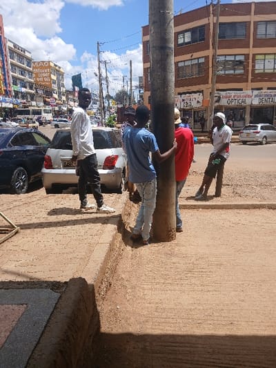 The image depicts an outdoor urban scene in Huruma ward, Kenya, during daylight under a partly cloudy sky. Five adult males are centrally located near a thick, dark utility pole. A dark-skinned male in a white long-sleeved shirt, dark trousers, and white sneakers stands on the left, facing the viewer. Another dark-skinned male, in a light blue t-shirt and faded jeans, is positioned by the pole, holding it with his left hand, his back mostly to the viewer. A third male in a red long-sleeved shirt is partially visible behind the second. A fourth male's head, wearing a dark cap, is visible between the first two. On the right, a fifth dark-skinned male wears a white and green short-sleeved shirt, dark shorts, and a white cap, leaning against the pole or an adjacent unseen object.

The immediate foreground features a reddish-brown textured concrete sidewalk with a raised, partially eroded curb. The street beyond hosts parked vehicles: a dark sedan and a silver hatchback (registration starting KBR). In the background, multi-story commercial buildings line the street. The building on the right has a reddish-brown facade and features "DELIGHT OPTICS LIMITED OPTICAL" and "DENTAL" signage on its ground floor. Buildings on the left display various signs, including a prominent 'MAO Eldoret order on Oil' billboard. Overhead, numerous utility lines are visible. The overall scene indicates a busy commercial street environment.