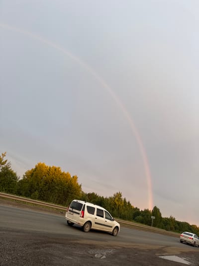 The image depicts a rural roadside scene in Sovkhoznyi, Russia, with a rainbow arching across a cloudy sky. The orientation of the image is vertical. The sky is overcast, suggesting a recent or ongoing rain shower, which has also left puddles on the asphalt road. Two white cars are visible on the road. The car in the foreground is a white hatchback, parked on the right side of the road. Further down the road, another white vehicle, possibly a van or SUV, is parked.
The road is flanked by a dense line of trees, with some trees showing vibrant yellow and green foliage, indicating it might be late summer or early autumn. A metal guardrail runs along the side of the road. A road sign is also visible amidst the trees. The overall mood is serene, with the rainbow adding a touch of natural beauty to the otherwise ordinary roadside setting. The time of day appears to be late afternoon or early evening, given the soft, diffused light. No people are visible in the image, and there is no discernible text.