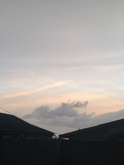 The image shows a view of the sky from a rooftop in Gaga, Nigeria, located at latitude 7.304 and longitude 5.153. The sky is a pale gray with a light pink hue on the horizon. There are a few white clouds in the sky, one of which is large and in the shape of a dragon. Two rooftops are visible in the foreground, both are silhouetted against the sky. There are no people in the image. The rooftops are both made of corrugated metal and are in a state of disrepair. There is a single electrical wire running from the left side of the image to the right.