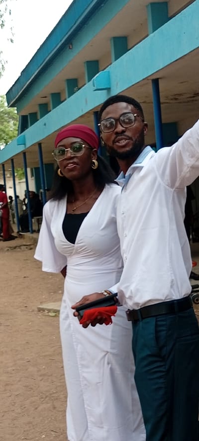 Two young adults pose for a photo in Goron Dutse, Nigeria. The woman wears a white dress, round sunglasses, gold hoop earrings, and a red headscarf. The man wears a white button-down shirt, black-rimmed glasses, dark trousers, and a black belt. The man raises one arm. The woman holds a black object with a red cloth. They are standing in front of a light blue building with pillars. Other people are visible in the background. The ground is dirt or sand.