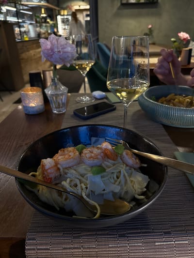 This image depicts a close-up view of a table setting in what appears to be a restaurant. The foreground is dominated by a dark bowl filled with pasta and several large shrimp, garnished with basil and shaved cheese. A gold fork and spoon rest on the rim of the bowl. To the left, a lit candle in an ornate holder provides a warm glow. Behind the pasta, a wine glass filled with white wine is visible, and further back, another glass of wine is also present. In the background, a pink peony sits in a small glass vase. Other restaurant patrons and staff are blurred in the distance. The overall ambiance suggests an evening meal in a dimly lit, upscale establishment. The setting is the restaurant "Holl's Bräustüberl" in Mayrhofen, Austria.