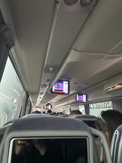 The image is taken from inside a bus, looking up towards the roof, with the rear of the seats and the back of the headrests visible. There are two small screens on the ceiling playing videos. The bus is likely moving as the outside window shows blurred scenery. The location, Guadarrama, Spain, at the coordinates 40.697, -4.127, is a town in the Sierra de Guadarrama mountain range, known for its natural beauty and hiking trails. It is possible this bus is part of a tour group or transporting local residents. The image does not reveal the exact bus route. There are several people sitting on the seats ahead, however, they are obscured from the viewer.