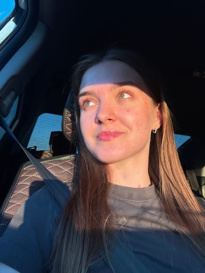 The image shows a young woman sitting in the driver's seat of a car, looking out the window. The car's interior is visible, including a seatbelt, quilted seat upholstery, and the dashboard. The woman has light-colored eyes, pink-tinted eyelashes, and is wearing a gray t-shirt and a small hoop earring. Sunlight is casting a warm glow on her face, highlighting her features and creating a shadow across her forehead and right cheek. The background outside the car window appears to be a landscape with a blue sky. The image does not provide any specific information about the location in the city of Ustinovo, Russia.