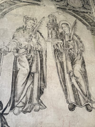 This is a fresco painting located in the atrium of the Convent of San Miguel Arcángel in Huejotzingo, Mexico. The painting depicts two female figures, likely saints or religious figures, rendered in a monochromatic palette of black and grey. They are depicted standing, facing slightly towards each other, and are elaborately robed in draped garments. Both figures wear halos, suggesting their religious significance. The figure on the left holds a palm frond and a cross-like object. The figure on the right holds a palm frond and a small building, possibly a church or bell tower, in her left hand, and a book in her right. The background is a plain, textured wall, consistent with a fresco medium, and above the figures, architectural elements of the fresco, such as arched structures and decorative motifs, are visible, also rendered in grey tones. The painting shows signs of age and wear, with visible cracks and fading in the plaster.