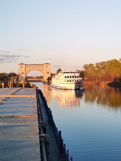 A riverboat passes under a stone archway in Myshkin, Russia. The river is calm, reflecting the boat and the sunset light. A concrete dock with mooring equipment is visible in the foreground. Trees line the riverbank opposite the dock.