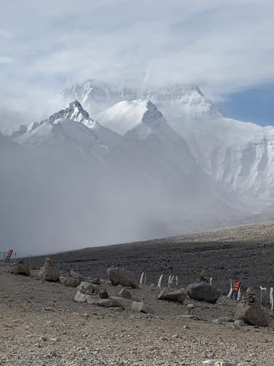 The image depicts a mountainous landscape in Tashi Dzom town, China. A large snow-covered mountain dominates the left side of the frame, partially obscured by clouds and mist. To the right of the mountain is a rocky, barren landscape. Small white flags and some flags with colored markings are interspersed with rocks along the ground. The composition is oriented in a vertical format. The sky visible above the mountain is pale blue.