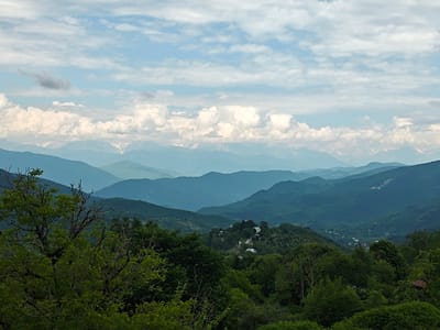 This is an eye-level, medium shot of a mountainous landscape in Ukeshi, Georgia. The sky is filled with scattered white clouds against a pale blue background. Several layers of mountains recede into the distance, with the furthest peaks appearing snow-capped and bathed in a hazy light. In the mid-ground, a village with white-roofed houses is nestled on a hillside amongst lush green trees. The foreground is dominated by the dense foliage of large trees and branches, partially obscuring the view of the village and the rolling hills. The overall impression is one of a serene, natural environment.