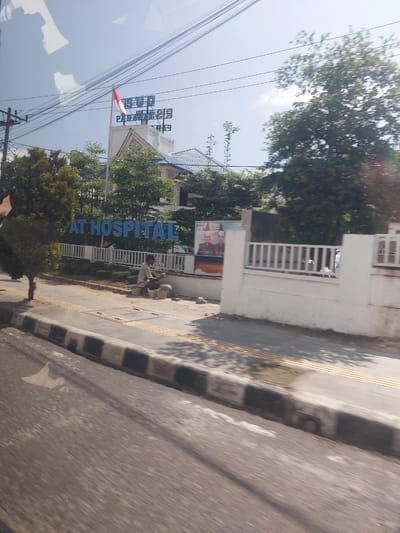 The image shows a view of a building with a sign that reads "AT HOSPITAL". A man is crouching on the sidewalk in front of the building, appearing to be working with some materials. There are trees and a fence surrounding the property. The sky is visible in the background, and there are power lines overhead. The scene appears to be in a city, possibly Parapat, Indonesia, given the context.