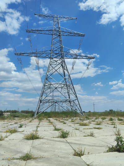 The image shows a large, latticed metal electricity pylon against a bright blue sky with scattered white clouds. The pylon is the dominant subject, seen from a low angle looking upwards. Wires extend from the top of the pylon, disappearing into the sky. The ground in the foreground is a flat expanse of concrete slabs, cracked and uneven, with patches of green grass and small yellow wildflowers growing in the gaps. In the distance, more electricity pylons are visible, stretching across a flat, grassy landscape with some trees and shrubs. The scene appears to be an open, somewhat neglected area, possibly on the outskirts of a settlement. The location is identified as Bilyaivka, Ukraine.