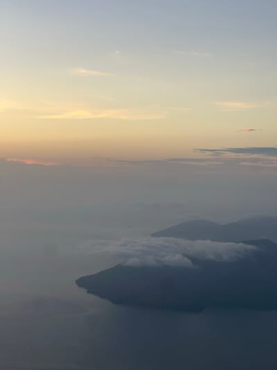 The image captures a serene aerial view of a body of water, possibly a lake or bay, adjacent to a mountainous landmass partially shrouded in mist. The sky displays a gradient of soft hues, transitioning from a pale blue at the top to a warm yellow and pinkish orange near the horizon, indicative of either sunrise or sunset. wispy clouds are scattered across the sky, with a more prominent bank of clouds clinging to the mountain peaks, softening their contours. The water surface is mostly calm, reflecting the muted colors of the sky. The overall impression is one of tranquility and natural beauty. The prompt specifically requests a description of El Cacao, El Salvador, however, no identifiable landmarks or specific details within the image allow for definitive location confirmation. The scene is devoid of people, specific objects, or actions.