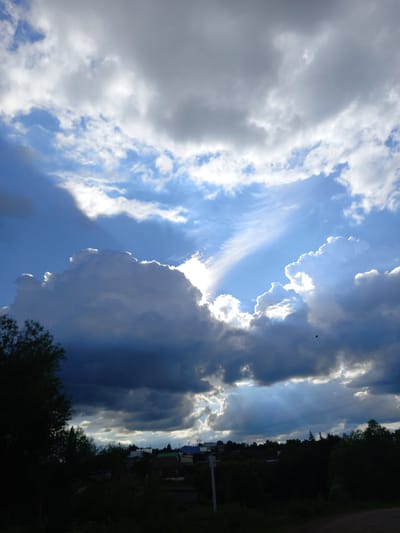 The image depicts a dramatic sky with a mix of light and dark clouds, suggesting a transition in weather. Sunlight breaks through the clouds, creating visible rays that illuminate the blue sky. The scene appears to be a rural or semi-rural setting, with hints of buildings and trees visible along the horizon, silhouetted against the bright sky. A single bird is visible as a small dark speck in the upper portion of the frame. The orientation of the image is vertical. The time of day is likely late afternoon or early evening, given the angle of the light and the dramatic cloud formations. The overall mood is one of awe and perhaps anticipation due to the powerful display of nature. There is no visible text in the image.