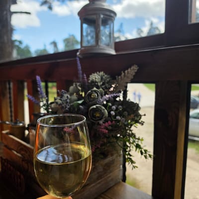An outdoor scene is depicted, with a glass of white wine prominently featured in the foreground. The glass is held by a person whose hand is partially visible at the bottom of the frame. Behind the wine glass, a wooden planter box contains an arrangement of artificial flowers and greenery, including purple lavender-like stalks and muted green roses.

In the background, a decorative lantern with glass panes is perched on a wooden railing. Beyond the railing, a blurred view of trees and a bright blue sky with scattered clouds is visible through what appears to be a window or open doorway. The context suggests a relaxed outdoor setting, potentially a cafe or restaurant with a view, possibly located in Atoluka, Bulgaria, given the implied context.