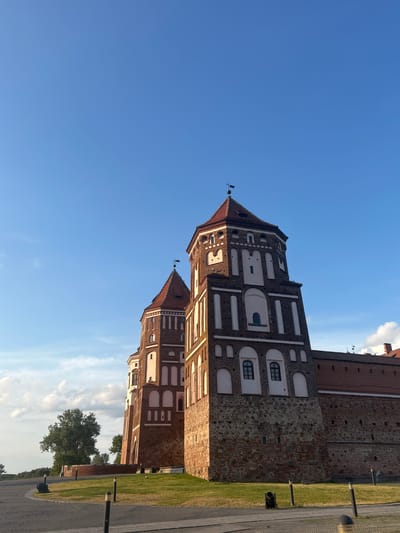 The image shows a view of Mir Castle, a UNESCO World Heritage site located in the settlement of Mir in the Karelichy District of Grodno Region, Belarus. The castle is a large, multi-towered fortress constructed from red brick and stone. The sky above is a clear blue with a few wispy clouds. In the foreground, a paved area leads up to a grassy slope that surrounds the castle walls. There are several decorative lampposts and a historical cannon positioned on the grounds. Trees are visible in the background.