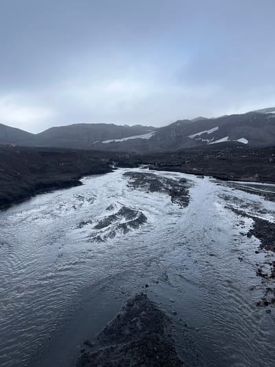 A gray, overcast day illuminates a landscape of stark contrasts. Dark, rugged mountains partially covered with snow rise beside a rapidly flowing, silty river. The river's surface reflects the diffused light, creating a shimmering effect. Gravel and debris line the riverbanks. The overall scene evokes a sense of cold, rugged terrain, and powerful natural forces.