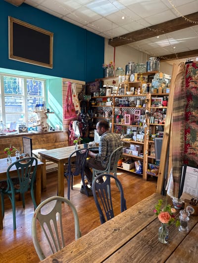 The image shows the interior of a cafe or shop, likely in West Oxfordshire, UK, given the visible street view. A man wearing a plaid shirt is seated at a wooden table, seemingly looking at something in front of him. The table is set with small vases of flowers, a menu, and condiments. Behind the man is a wall of shelves filled with an assortment of items, suggesting a gift shop component to the establishment. A window is visible with light flooding in, and a blackboard hangs on the blue wall in the corner. The chairs are a mix of colors and designs. The ceiling has exposed beams and string lights.