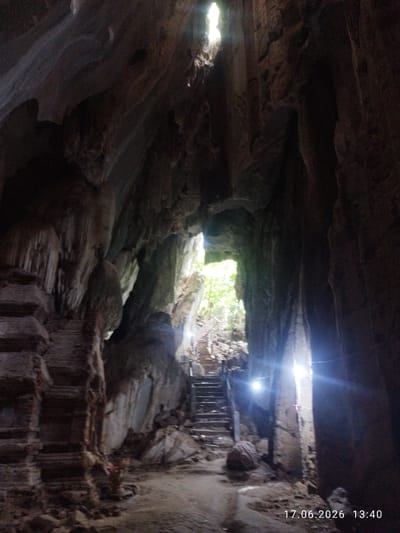 A cave interior with a natural light opening at the top, illuminating a set of stairs leading upwards. The cave walls are rough and rocky, with formations that resemble stalactites and stalagmites. To the left, a tiered structure, possibly ancient steps or a platform, is visible. The ground is uneven, covered with dirt and rocks. Artificial lights are present within the cave, casting illumination. The overall impression is of an ancient and natural environment, likely a tourist attraction or a place of historical significance. A date and time stamp "17.06.2026 13:40" is superimposed on the bottom right corner of the image.