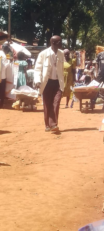 The image captures an outdoor market scene in Shirungu-Mugai ward, Kenya, under bright daylight. The ground is unpaved, consisting of reddish-brown dirt with scattered small debris.
In the foreground and center, an adult bald male, appearing to be of African descent, walks from left to right. He is dressed in a light-colored, possibly off-white, collared jacket over a white long-sleeved shirt, dark brown trousers, and dark shoes. A silver-colored crucifix on a chain is visible over his shirt. His head is turned slightly to his right.
To the left of the central figure, a person in white attire and a young child wearing a light green dress are partially visible. Adjacent to them is a low-standing vendor's stall or cart, with items covered by a white cloth. To the right of the central figure, a woman in a patterned yellow and green dress is partially visible behind him. Further right, another market stall displays various textiles or clothing items. A person wearing a dark shirt with a visible "Fly Emirates" logo is also seen in the background on the right. Additional individuals are present throughout the mid-ground and background, indicating a populated public or commercial area. Large green-leafed trees form the distant background. Shadows are cast on the dirt ground, consistent with overhead sunlight.