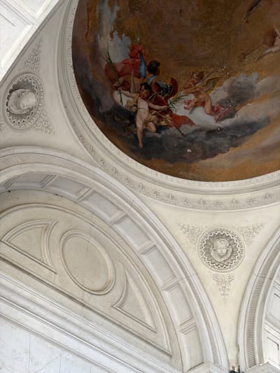 The image shows a fresco on the ceiling of a domed structure. The fresco depicts several cherubic figures in flight amidst clouds. One cherub is prominently positioned in the center, holding a branch and an arrow. Other cherubs are seen around it, some with wings and others seemingly holding weapons or banners. The fresco's colors are rich, with shades of brown, red, blue, and flesh tones dominating.
Below the fresco, the architectural elements of the dome are visible. These include plasterwork with ornate moldings and carved decorative motifs, such as rosettes and stylized floral patterns. The architecture features curved arches and recessed panels, suggesting a classical or Baroque style. The overall impression is one of grandeur and artistic embellishment.
The image's metadata indicates that the location is Mozzo, Italy. More specifically, this fresco is located within the Santuario della Madonna delle Lacrime in Mozzo, a sanctuary dedicated to the Virgin Mary.