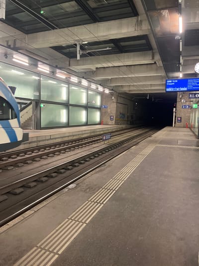 The image depicts an underground train station platform, consistent with a location in Chêne-Bourg, Switzerland. The perspective is tilted, showing the grey platform on the right and multiple train tracks on the left, receding into a dark tunnel. The front section of a blue and white train is partially visible in the bottom-left corner, adjacent to the tracks.
The station features concrete walls with large rectangular glass panels that reflect artificial light from overhead fixtures. The platform has tactile paving, marked by white striped patterns, running along its edge.
Key objects include a digital display board showing "DEC 19:39" and "Coppet" as a destination, with a list of stations beneath: "Genève-Eaux-Vives," "Genève-Champel," "Lancy-Bachet," and "Lancy-Pont-Rouge." A clock on the upper left wall also displays approximately 7:39 PM, indicating the evening. Other signs include platform or track identifiers like "2 A" and "A 1," along with pictograms for information, accessibility (wheelchair), and stairs/escalators.
The scene captures an operational train station, likely with a train stopped or in transit. The absence of people creates a calm, functional atmosphere in this modern, industrially designed setting.