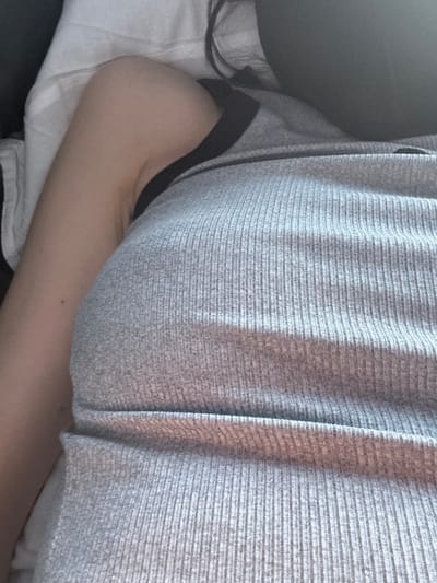 The image depicts a close-up, top-down view of a single individual in a resting or reclining position. The scene is located in Fetino, Russia. The subject's upper torso and left arm are prominently featured. The individual wears a light gray, ribbed tank top or camisole with a black trim along the left armhole/strap. The skin of the left arm is visible, appearing light in tone, with a small, dark mole located on its outer aspect, approximately midway along the visible portion. A segment of dark hair is discernible in the upper right portion of the frame. The left arm rests on a white or off-white textile surface, likely a pillow or bedding. A dark, indistinct surface, possibly upholstery, is visible behind the individual's head/hair area. No active interactions are occurring. The lighting is diffused, casting subtle shadows and highlights on the ribbed fabric of the garment.