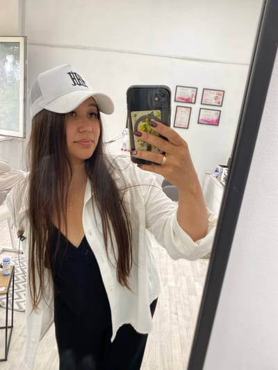 In a well-lit indoor setting, possibly a salon or studio, a woman is taking a mirror selfie. She is wearing a white trucker hat with a black logo and an open white shirt over a black tank top. Her long, dark hair is down, and she is wearing a gold necklace with a small pendant. On her right hand, she has a gold ring and dark nail polish. The mirror reflects the room behind her, which includes framed certificates on the wall and what appears to be a white cabinet. The floor is light wood laminate. The woman is positioned in the foreground, holding her black smartphone to take the picture. The overall impression is one of casual self-documentation within a professional or personal grooming space. The location is specified as Akyar, Russia, though the image itself does not provide direct visual evidence of this specific location.