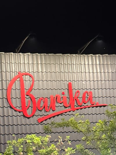 The image shows an exterior shot of a building, possibly a business, in Lambaro, Indonesia. The building has a gray corrugated metal wall with red signage spelling out "Bunker" in a cursive font. Some green foliage partially obscures the sign and wall in the upper right corner. A security light fixture is visible on the left edge of the wall against a dark background. The overall lighting suggests it is nighttime.