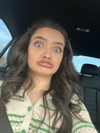 A young woman with long, wavy brown hair and light skin is in the driver's seat of a car. She is wearing a cream-colored sweater with green stripes and buttons. Her eyes are wide and her lips are puckered in a playful expression. The interior of the car is black. Through the car window, one can see a hint of sky and trees, suggesting an outdoor setting. It is impossible to discern the exact location in Zagorsko, Bulgaria, from this image.