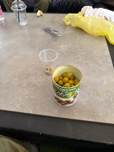 The image shows a grey table top with a can of green olives, a plastic cup with a few nuts in it, and an empty plastic cup with a straw sticking out of it. The table is located at the coordinates 31.833 latitude and 34.82 longitude, in Mazkeret Batya, Israel. The table is made of smooth, flat, grey tiles and has a rough texture. There is a yellow plastic bag on the table, folded on the upper right side of the image. The can of olives is in the center of the image, open and filled with green olives. The olives are arranged in a random pattern inside the can. The can is labeled in Hebrew and English, with "Green Olives" visible in English. The plastic cup with the nuts is on the left side of the can. It is a clear plastic cup with a white lip, and the nuts are brown and scattered at the bottom. The other plastic cup is on the left side of the image, empty and with a straw protruding out of it. The straw is black and white. The image is taken from a high angle, looking down at the table. The background is out of focus and shows a dark color.