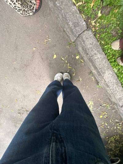The image shows a low-angle perspective of a person's legs and feet standing on a paved surface, possibly a sidewalk or walkway. The person is wearing dark denim jeans and white athletic shoes. To the left of the frame, another person's leg is visible, wearing leopard-print pants and a sandal with bright red nail polish. The ground is a mixture of asphalt and some scattered dry leaves. To the right of the paved surface, there is a strip of green grass growing next to a concrete curb that appears to be cracked. Another person's feet are visible in the grass area, wearing light-colored shoes and dark pants. The scene suggests an outdoor urban environment, possibly a park or street. The location is identified as Vyshchi Vil'shany, Ukraine.