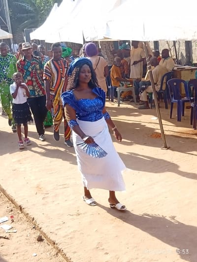 The scene is an outdoor gathering in Isinyi-Nando, Nigeria, captured on 19/12/2025 at 15:53. The primary subject is a dark-skinned woman walking forward on a sunlit, unpaved dirt path, positioned slightly right of center. She wears a vibrant blue embroidered sleeveless top with a matching blue headwrap and a white, eyelet-detailed full-length skirt. She holds a folded blue fan in her right hand and wears white open-toed sandals. Behind her, a group of people walks in a loose procession. This group includes a child in a white shirt and dark skirt, an adult male in a green patterned outfit, and several other individuals in various colorful traditional Nigerian garments. In the background, multiple white canopy tents are erected, under which numerous people are gathered. Some individuals are seated on blue plastic chairs around tables, while others stand. The ground throughout the visible area is light brown dirt. Strong sunlight casts distinct, long shadows, indicating an open-air environment. Minor debris, including small pieces of paper and a red bottle cap, are visible on the ground in the lower left foreground.