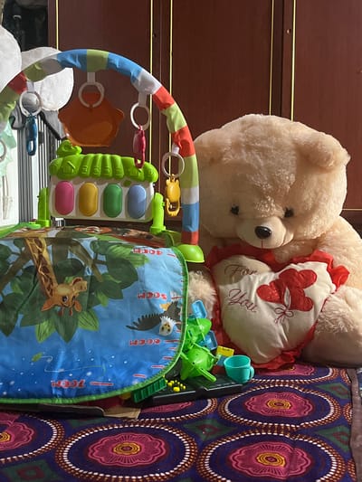 In the city of Kuyun, Uzbekistan, a plush teddy bear sits on a patterned rug, holding a heart-shaped pillow embroidered with "For You" and two red balloons. Beside the bear is a baby activity gym with hanging toys, a keyboard with colorful keys, and a play mat featuring a giraffe and other jungle animals. Several small toys, including a green plastic object and a teal cup, are scattered around the play area. The scene appears to be indoors, with a wooden door visible in the background.