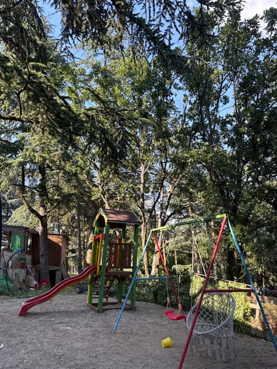 The image depicts a children's playground nestled within a dense forest, likely located in the city of Pobeda, Bulgaria. The playground features a red and yellow slide attached to a wooden play structure with green supports, and a swing set with both a red seat and a white macrame hanging chair. A yellow bucket is visible on the sandy ground near the swing set. The surrounding environment is dominated by tall trees with lush green foliage, creating a shaded and natural atmosphere. In the background, to the left, a wooden structure resembling a small house or shed is visible. The ground is covered in sand and small pebbles. The scene is peaceful and appears to be unoccupied.