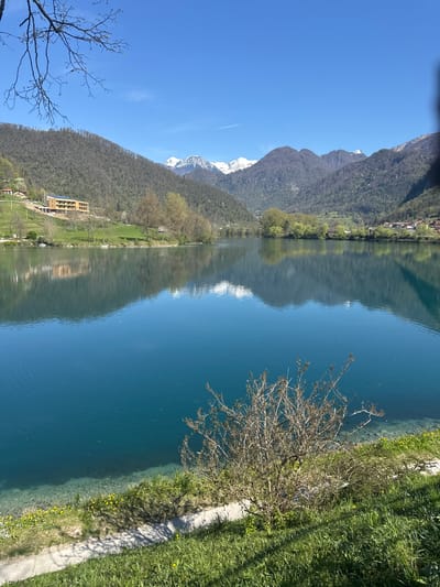 This image captures a scenic view of Lake Bohinj in Tolmin, Slovenia, on a sunny day. The clear blue water of the lake reflects the surrounding mountains, some of which are still capped with snow. The shoreline is lined with lush green vegetation, including trees, grass, and bushes, along with a paved walkway. The image also features a multi-story building with multiple windows at the base of a steep hill/mountain, suggesting a combination of natural beauty and development in the area.