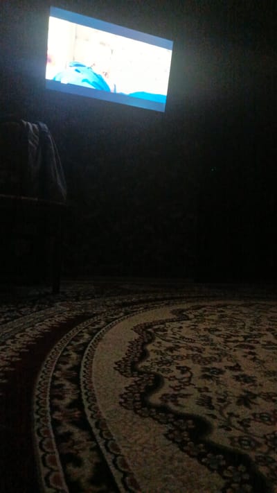 The image captures a dimly lit room, likely a living space, with a projector screen displaying a bright image in the upper left quadrant. The screen shows a close-up of a person wearing a blue garment, with a blurred background. In the foreground, a patterned carpet covers the floor, with its intricate design visible under the ambient light. To the left, a dark piece of furniture, possibly a sofa or chair, is partially visible, with a draped cloth on top. The overall ambiance is dark and quiet, suggesting a setting for viewing a movie or presentation. The location is stated to be in Shamsiabad, Uzbekistan.