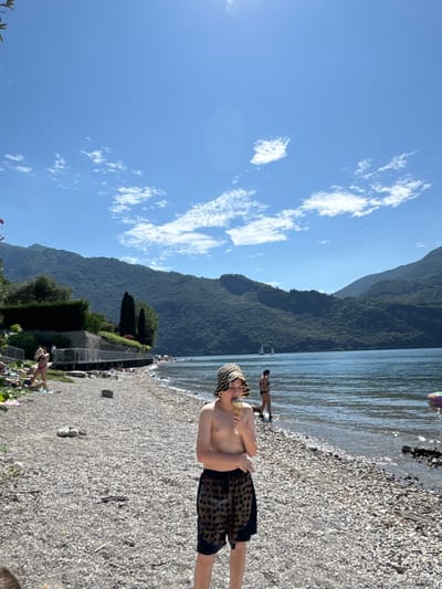 A sunny lakeside view features a boy in patterned swim trunks and a matching head covering, standing on a rocky beach while eating an ice cream cone. The water stretches towards a distant, forested mountain range under a blue sky dotted with fluffy clouds. In the background, other people are enjoying the water and beach. A retaining wall with greenery adds to the scene. The photograph is oriented sideways.