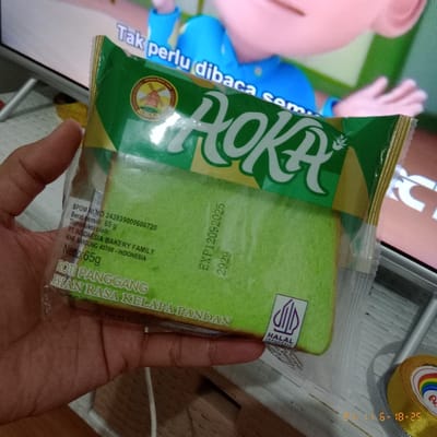 A hand holds a package of "AOKA" roti panggang (toast) with kelapa pandan (coconut pandan) flavor. The green and gold packaging displays the brand name in a stylized font, along with an image of a windmill. The package indicates a net weight of 65g and an expiration date of 12/09/2025. The product is halal-certified and manufactured by PT. Indonesia Bakery Family in Bandung, Indonesia. A television screen showing a cartoon character is blurred in the background.