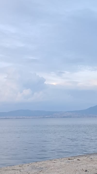 The image depicts a serene coastal scene in Νέα Μηχανιώνα, Greece. The foreground is occupied by a textured, light-colored surface, possibly a walkway or embankment, with scattered debris. Beyond this, a calm, expansive body of water stretches to the horizon. In the distance, a hazy coastline is visible, featuring rolling hills or mountains with subtle signs of a settlement or town. The sky above is overcast with soft, diffused clouds, with a faint glow of sunlight peeking through a gap in the clouds, suggesting either dawn or dusk. There are no people or animals visible in the frame.