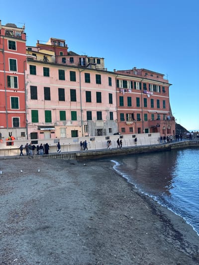 The image displays the waterfront of Vernazza, Italy, under a clear blue sky. A series of multi-story buildings, painted in red, pink, and terracotta hues, line the left side of the frame. The buildings feature numerous windows, predominantly fitted with closed dark green shutters. One pink building exhibits areas of peeling paint, and a terracotta building has several items of laundry drying on lines suspended from its upper facade. A white-lettered "GELATERIA" sign is visible on a shopfront in one of the lower floors of the pink building.
A light-colored concrete promenade, approximately 2.5 meters wide, runs along the base of these structures, separating them from a grayish-dark beach composed of sand and small pebbles. The promenade gradually inclines upwards towards the right of the frame. Approximately twenty-five individuals are observed walking or standing along this promenade. They are dressed in various casual outerwear, including jackets and coats, suggesting cool weather.
To the right, the deep blue water of the Ligurian Sea meets the beach, forming a distinct waterline with small, breaking waves. The water reflects the buildings to some extent. A small orange life ring is attached to a low wall section at the far left of the promenade. The perspective is slightly elevated, looking down towards the beach and across at the buildings and the activity on the promenade.
