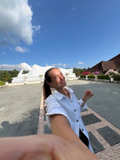 A woman, appearing to be in her late 20s or early 30s, is taking a selfie outdoors in front of a large white reclining Buddha statue. She has long, dark brown hair pulled back in a ponytail and is wearing a white, button-up shirt. She is smiling at the camera and her right hand is partially visible, reaching towards it.

The Buddha statue is enormous and is positioned on a raised platform. Behind the statue, there are lush green trees and a blue sky with scattered white clouds. To the right of the statue, there is a building with a reddish-brown tile roof and a row of flowering plants in white planters. The ground in the foreground is paved with large gray tiles and interspersed with gravel pathways. The scene appears to be a serene outdoor temple or garden complex. The location is identified as the Pupuan Buddha Temple in Pupuan, Bali, Indonesia.